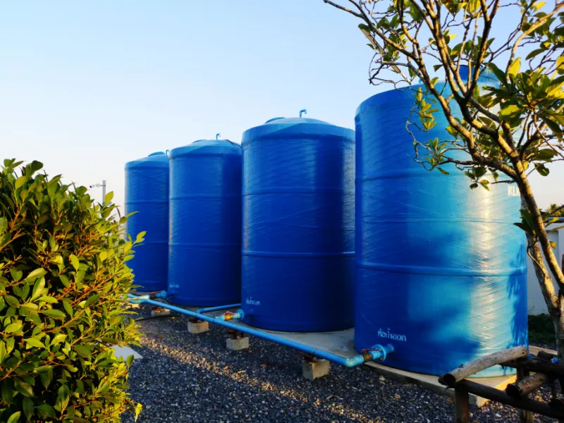 Large blue water storage tanks installed in a row outdoors on a gravel base with connecting plumbing pipes.